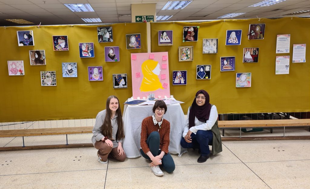 Amina MWRC CW How It Felt Team photo with 3 women standing in front of canvases with artwork of Muslim women wearing headscarves.