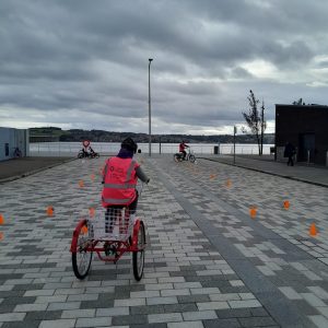 cloudy sky, paved ground, looking at back of a woman on a trike wearing a pink hi-vis jacket
